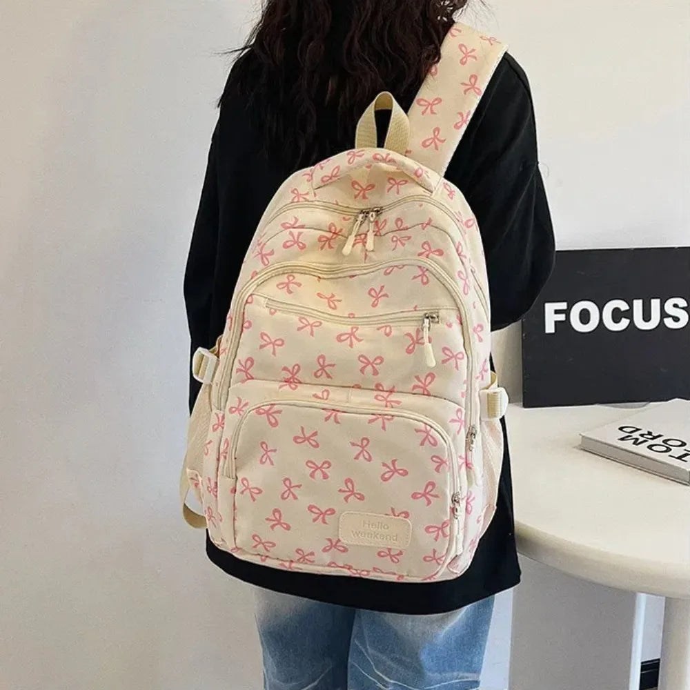 A cream-colored nylon backpack with pink bow prints is carried by a person against a white background.