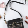 Un sac à main en cuir noir avec un rallonge de chaîne dorée et un fermoir en forme de diamant, exposé sur une surface blanche