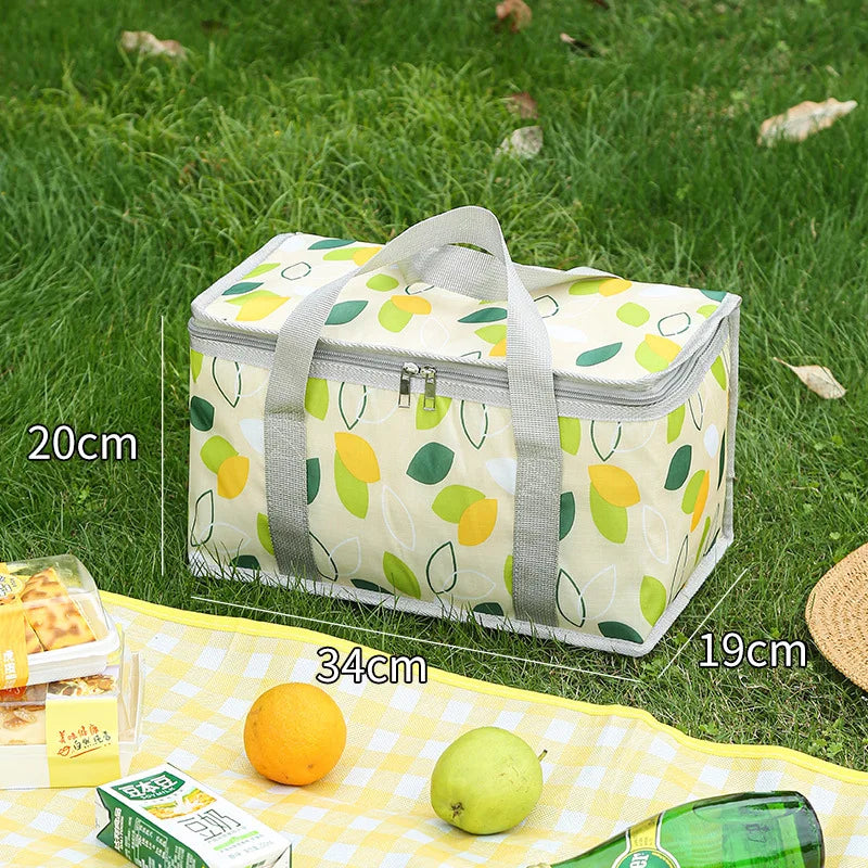 A white insulated picnic tote with a yellow and green leaf pattern is shown on grass next to citrus fruits and a bottle.
