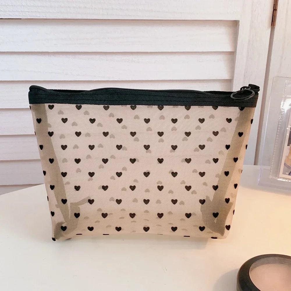 A beige mesh cosmetic storage pouch with a black trim and small black hearts pattern, displayed on a white surface.