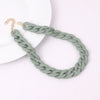 A close-up of a trendy acrylic statement choker necklace in a sage green chain resting on a white surface.
