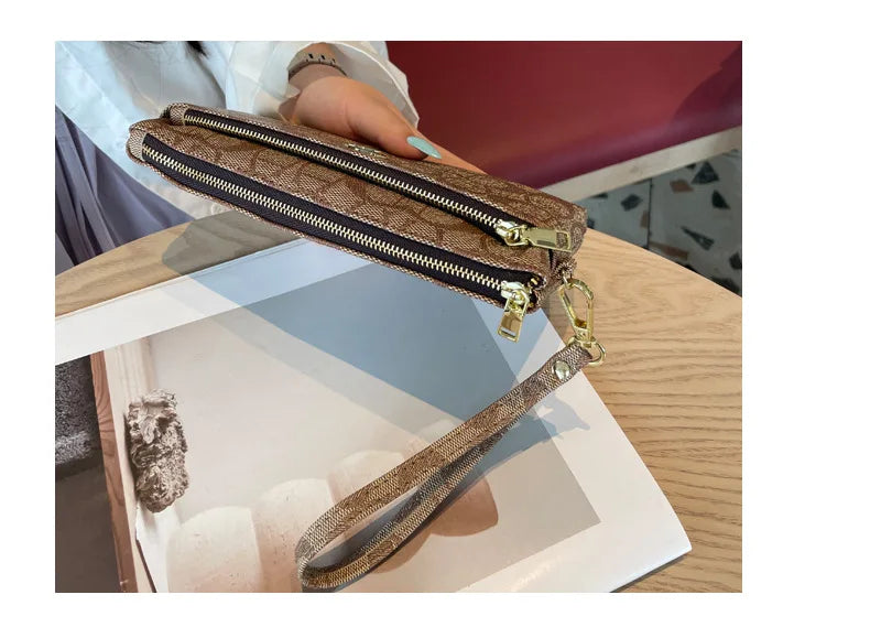 A brown textured zippered wallet with a gold-toned strap is shown being held by a hand on a wooden surface.