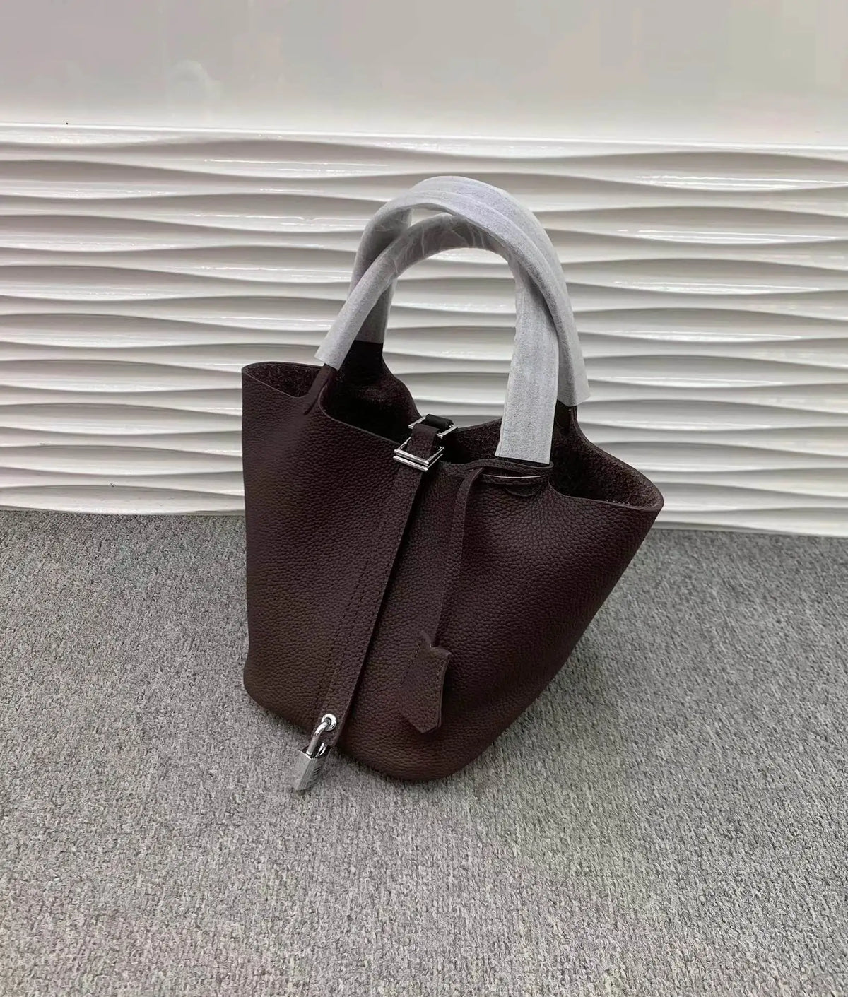 Dunkelbraunes Leder-Bucket-Bag mit hellem grauem Stoffgriff auf grauem Untergrund.