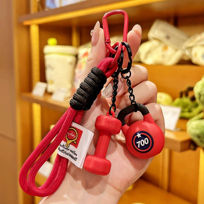 A hand holds a red nylon D-shape fitness keychain with a black cord and a red weight, set against a blurred background of gym