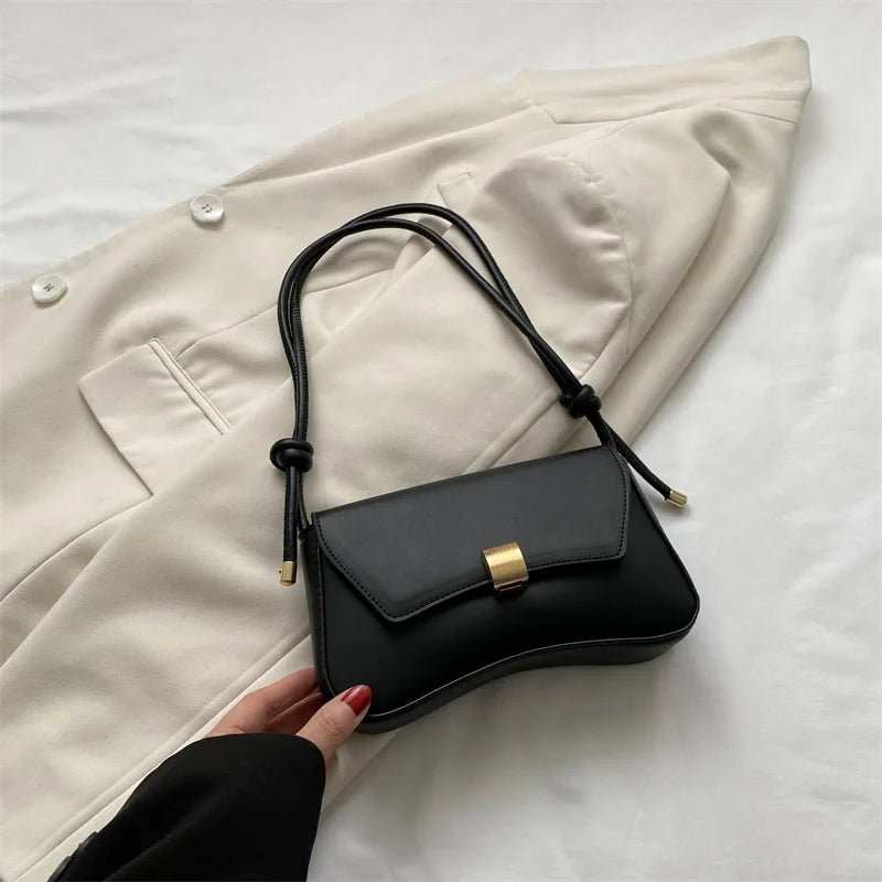 A black leather crossbody bag is held by a hand against a light beige blazer.