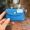 A hand holds a bright blue genuine leather card holder with a gold stud and a QR code.