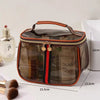 A transparent cosmetic bag with an orange trim and a handle, next to a glass bottle and makeup brushes.