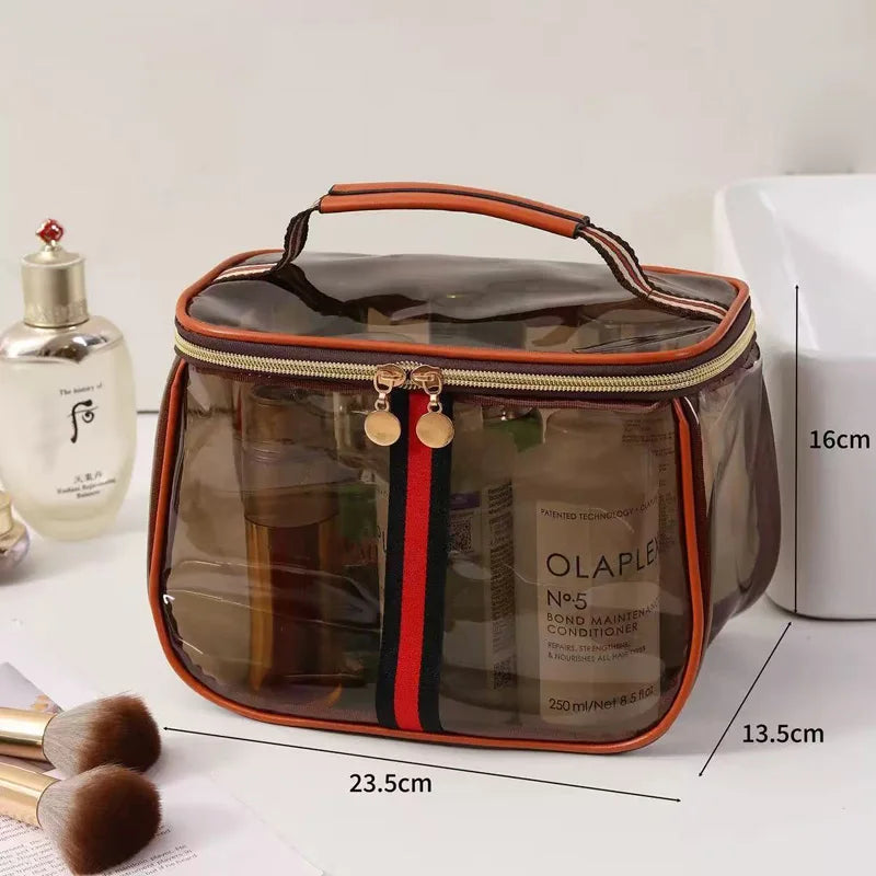 A transparent cosmetic bag with an orange trim and a handle, next to a glass bottle and makeup brushes.