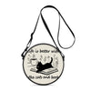 A round black cartoon cat crossbody bag with a leather strap and an illustration of a cat reading a book.