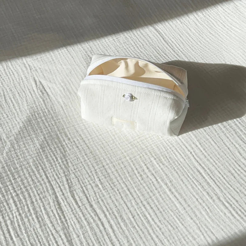 A white cotton toiletry pouch with a zipper is lying on a light striped fabric surface.