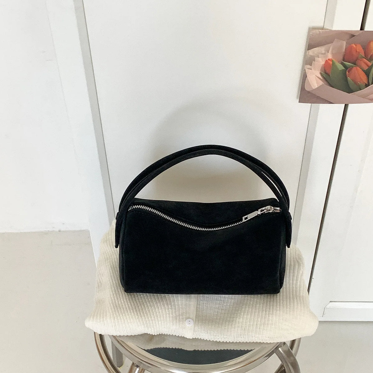 A black faux suede crossbody bag resting on a white knitted surface.