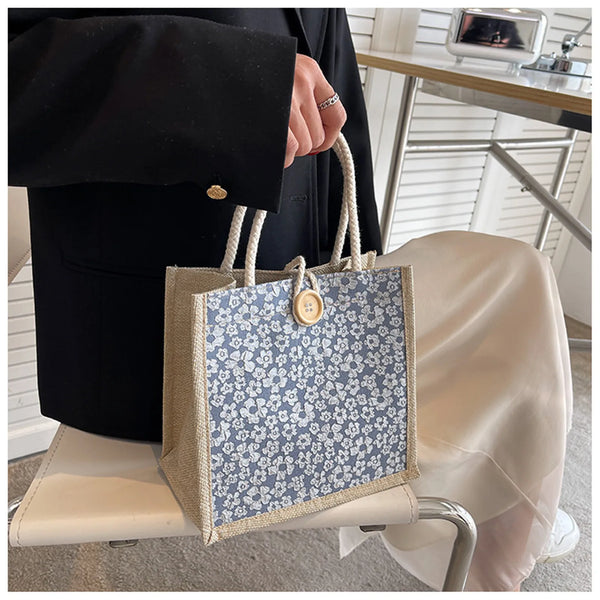 A beige floral linen tote bag with blue and white pattern is held by a person in a black suit jacket.