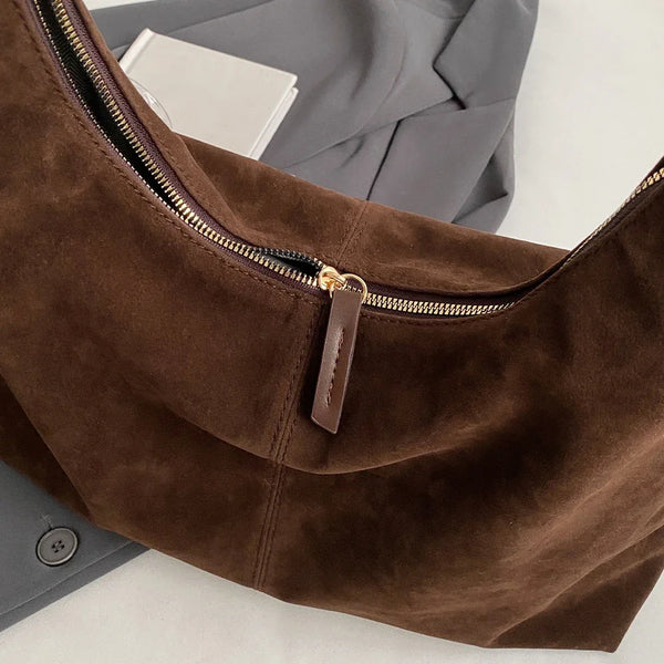 A close-up of a brown suede slouchy shoulder bag with a gold zipper and leather tab.