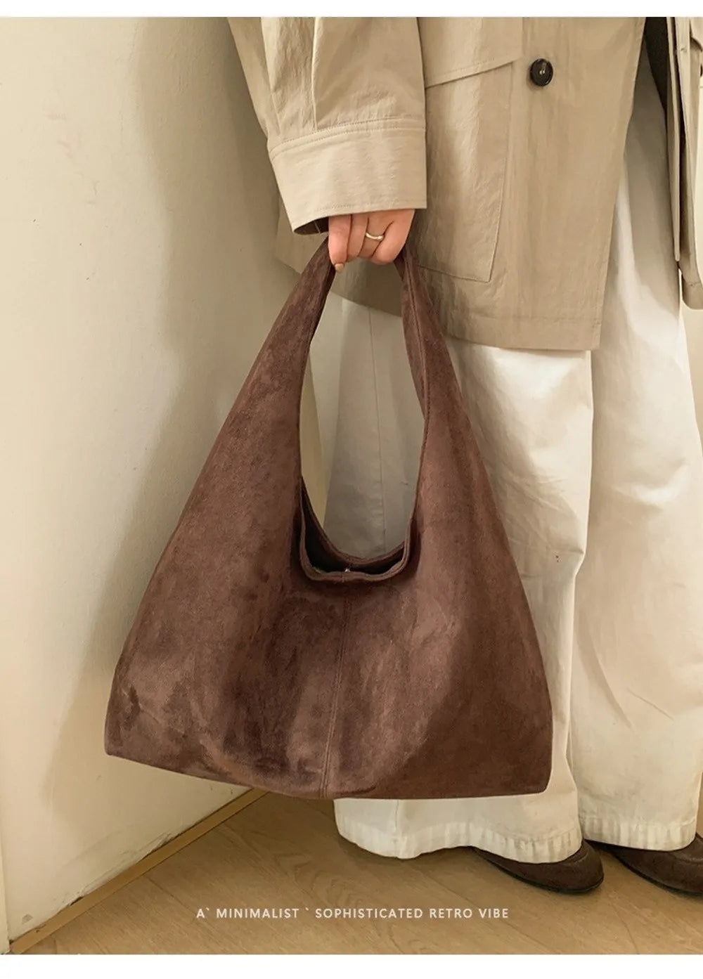 A brown suede shoulder tote bag being held by a person wearing light-colored pants.