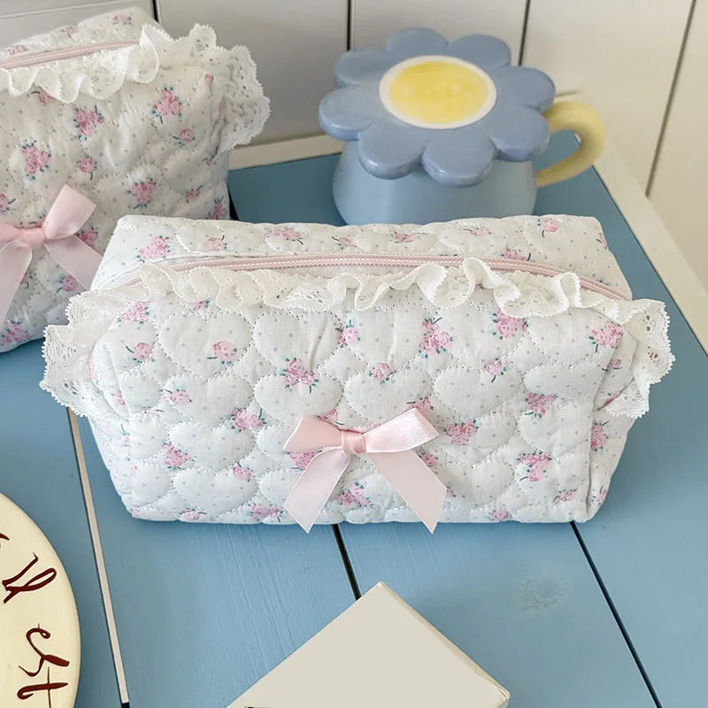 A white quilted cosmetic storage pouch with pink floral embroidery and a pink ribbon, next to a light blue ceramic cup.