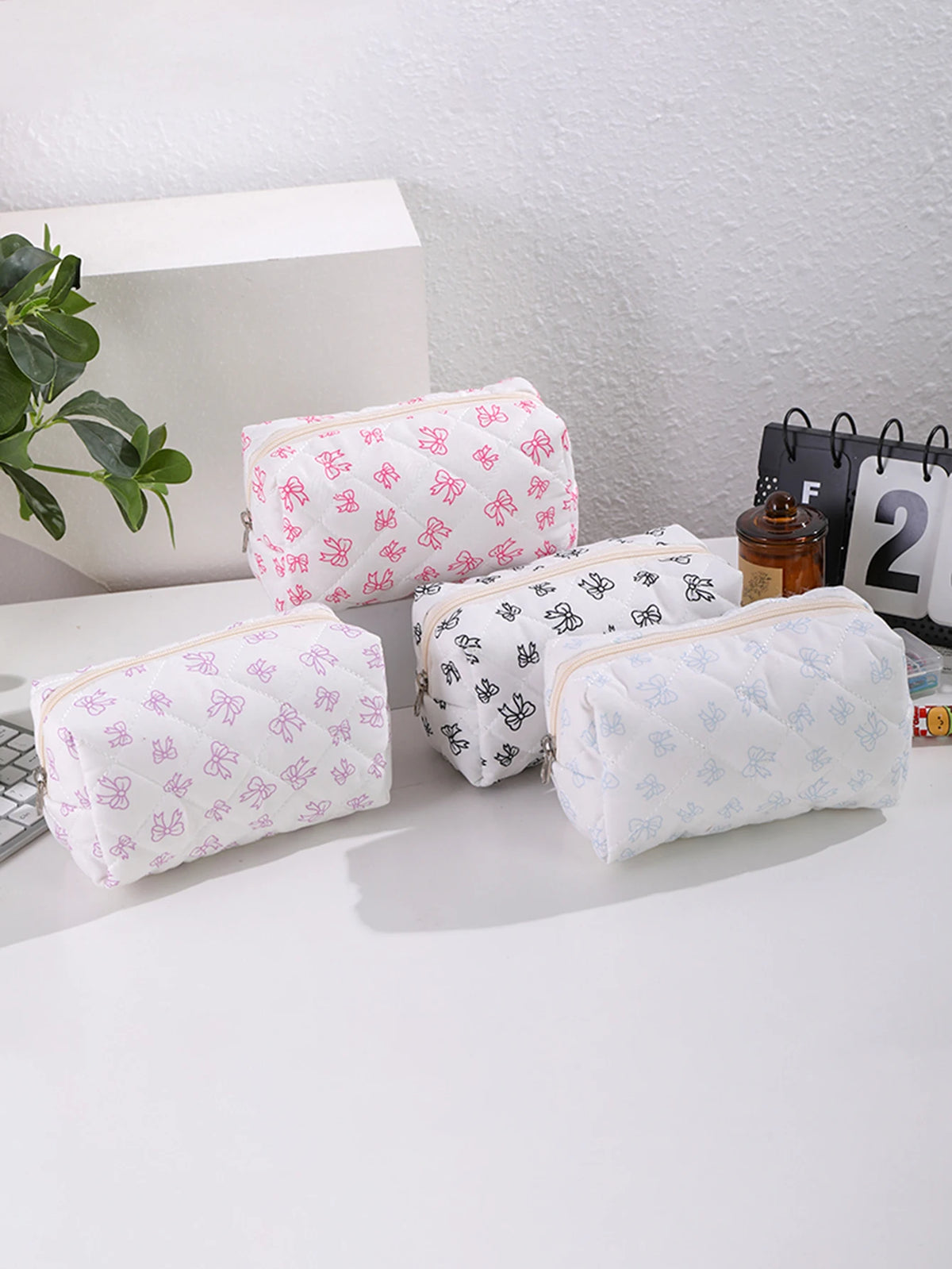 Una colección de fundas acolchadas de maquillaje rosa claro y blanco con estampado de lazo repetitivo, exhibidas sobre un sop