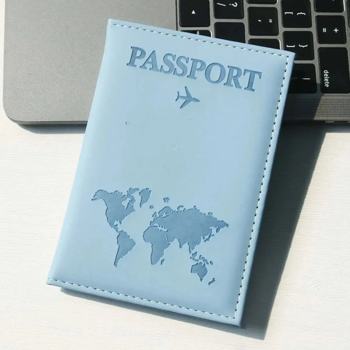 A light blue leather passport holder with a world map design is placed next to a silver laptop and a black keyboard.