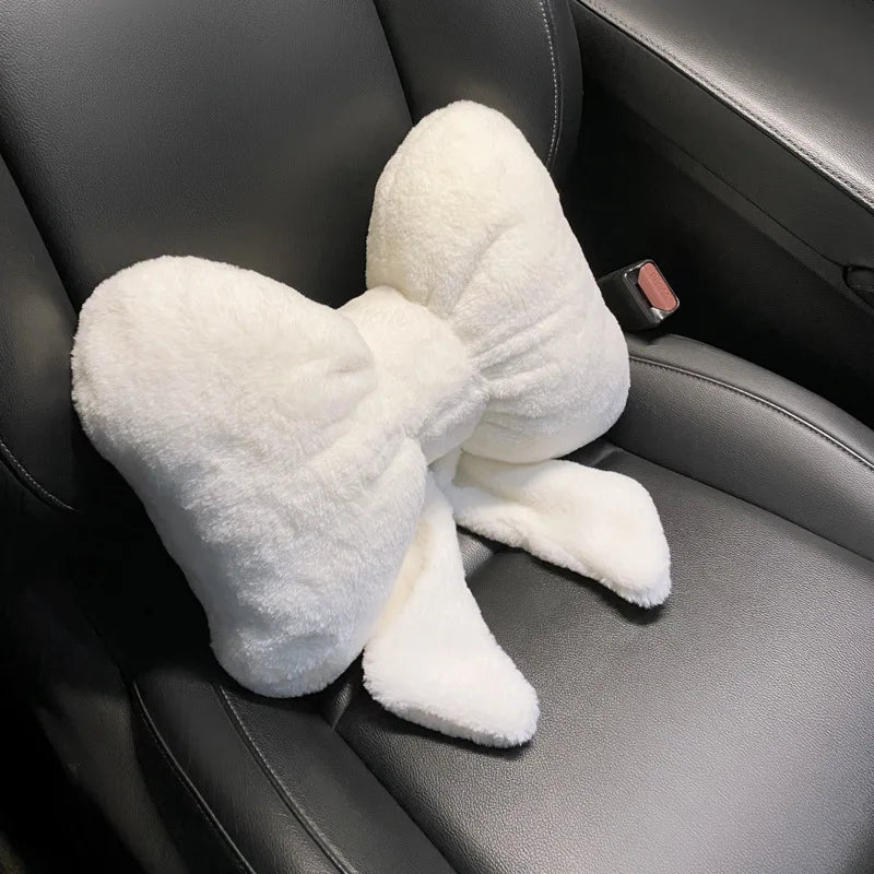 A white, fluffy bowknot car headrest pillow with lumbar support resting on a dark gray leather seat.