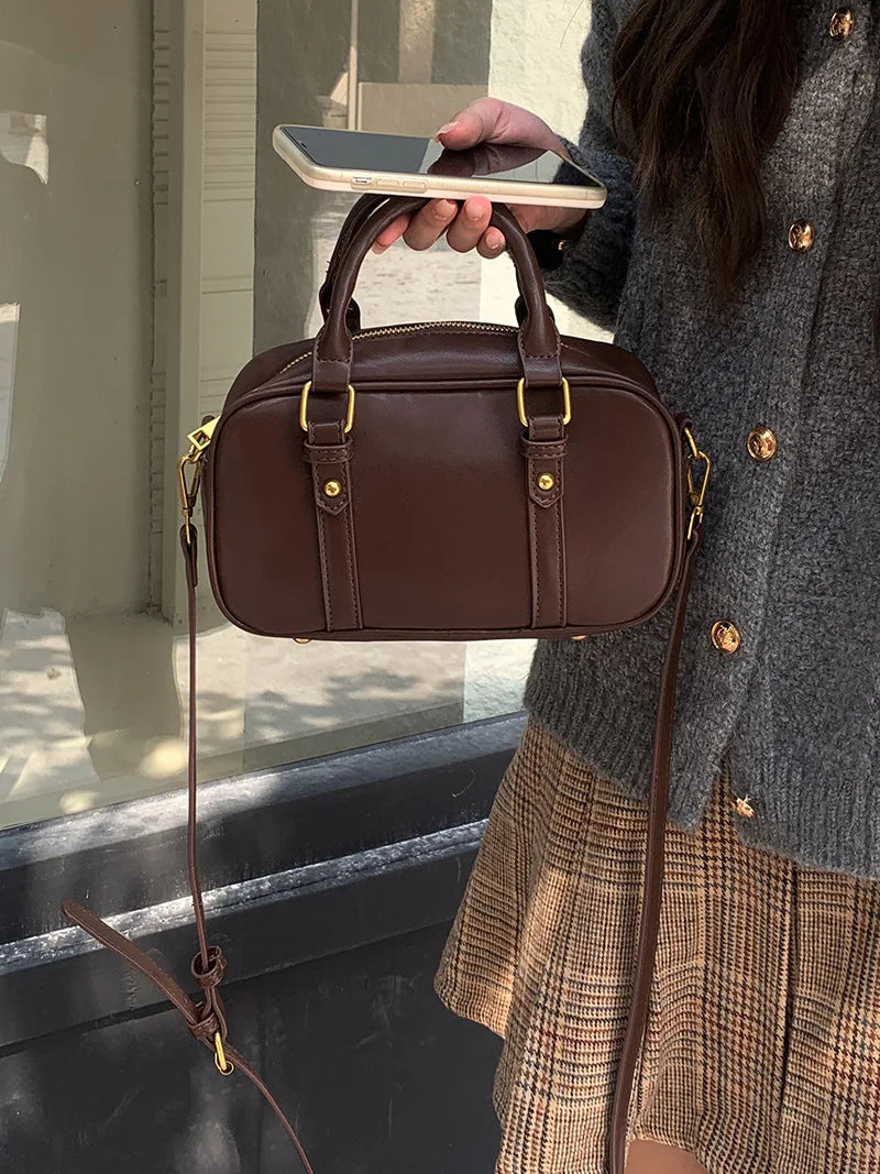 A brown vintage PU leather shoulder bag with gold hardware being held by a person against a light background.