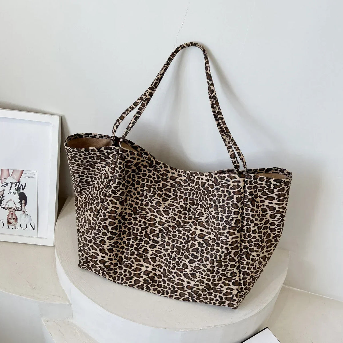 A brown leopard print canvas crossbody tote bag hanging on a white pedestal.