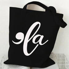 Black canvas tote bag featuring a white script design of 'la' with a ghost and pumpkin motif.