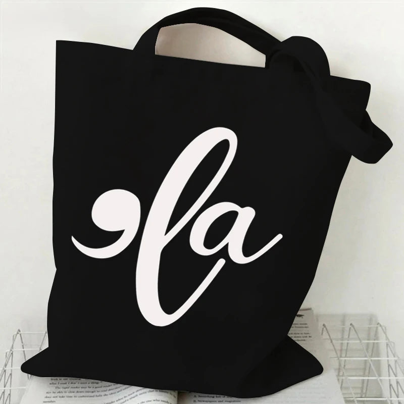 Black canvas tote bag featuring a white script design of 'la' with a ghost and pumpkin motif.