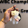 A white plush bear keychain featuring a camera and glasses is held in a person's hand against a blurred background.
