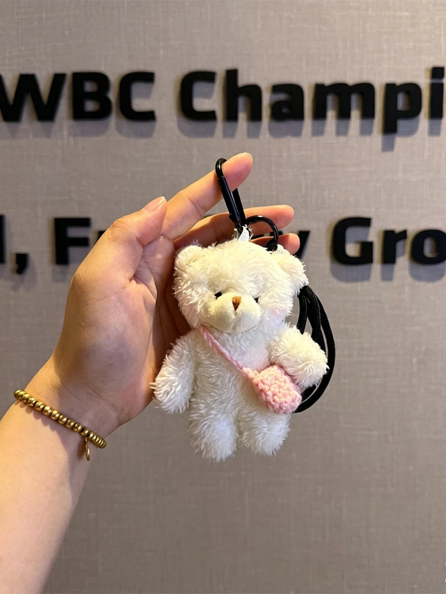A white plush bear keychain featuring a camera and glasses is held in a person's hand against a blurred background.