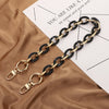 Une paire de bretelles de sac en acrylique, or et noir, détachables, posées sur un tissu brun à côté d'un papier blanc.