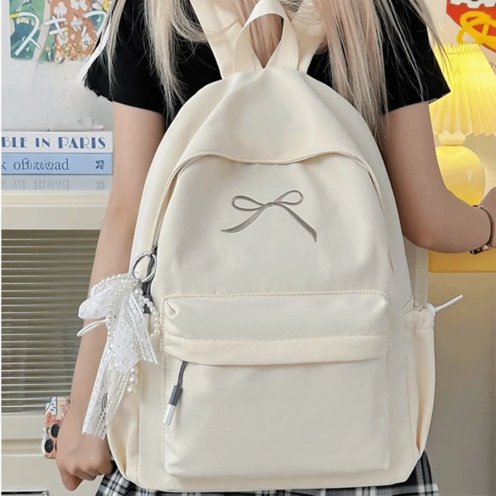 A light beige backpack with a bow detail and white lace trim is worn by a person.