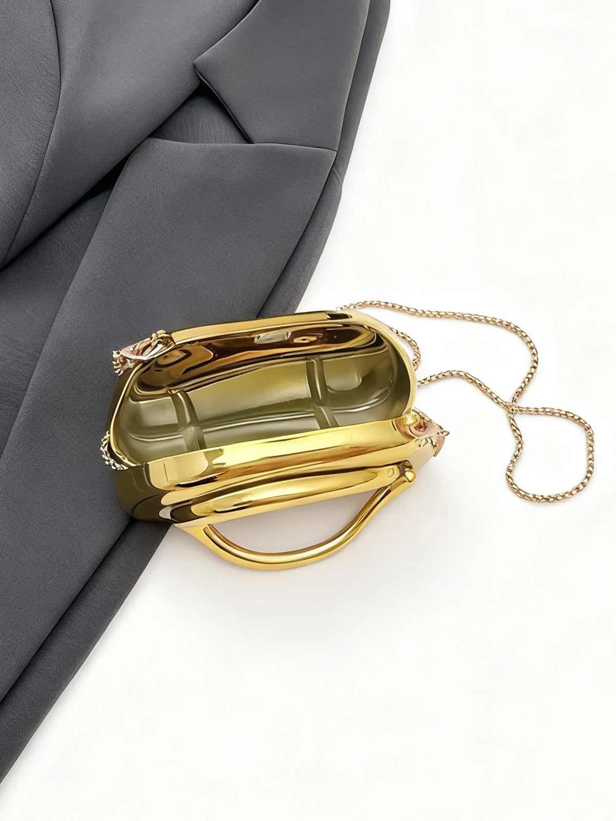 A small gold metal clutch bag with a gold chain strap is shown against a gray fabric background.