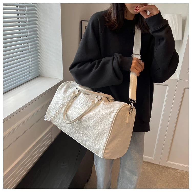 A woman holds a cream-colored crocodile grain PU leather travel duffle bag with a beige strap in a bright room.