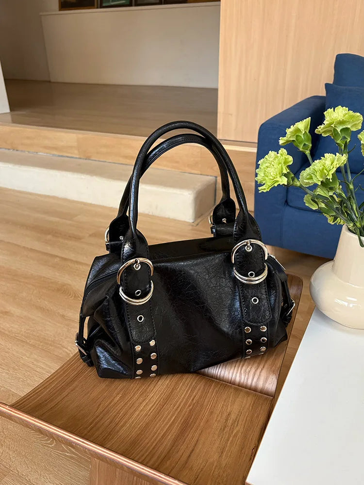 A black PU leather shoulder bag with silver rivets resting on a wooden surface next to a white vase of green flowers.