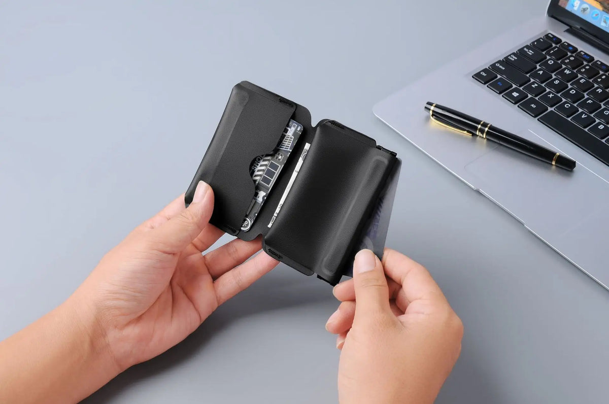 A hand holds a black magnetic foldable ID card holder and coin purse next to a silver pen on a laptop.