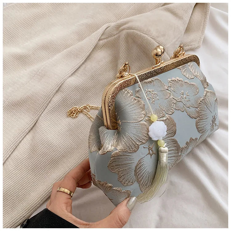 A light blue silk evening clutch with gold embroidery and a tassel charm is held against a beige fabric.