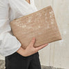 A beige textured clutch bag with a subtle alligator print is held by a person wearing a white shirt and black pants.
