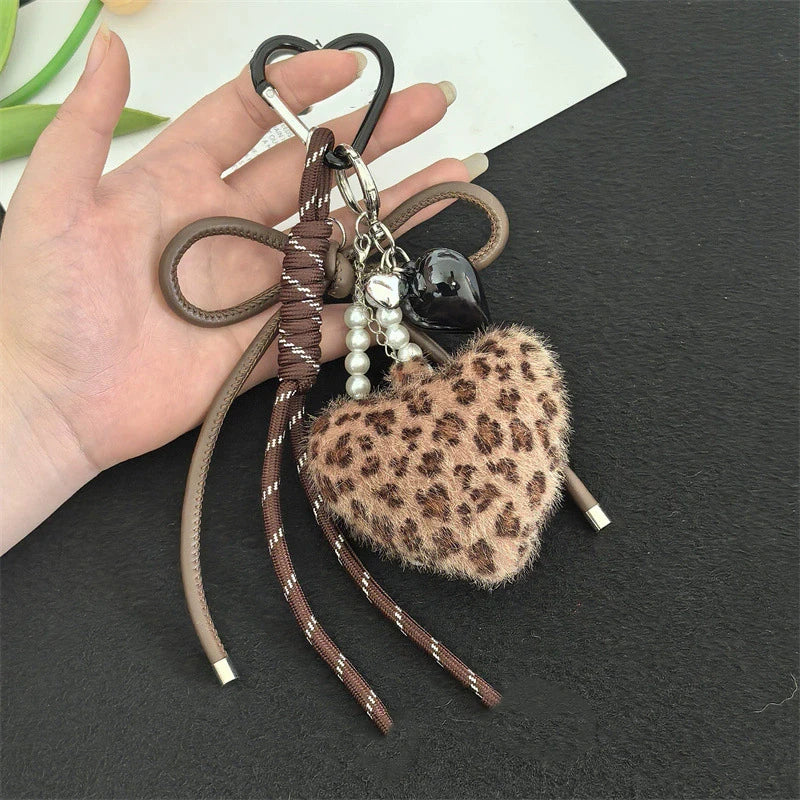 A leopard print faux fur charm keychain with brown leather straps and pearl beads is held in a hand.