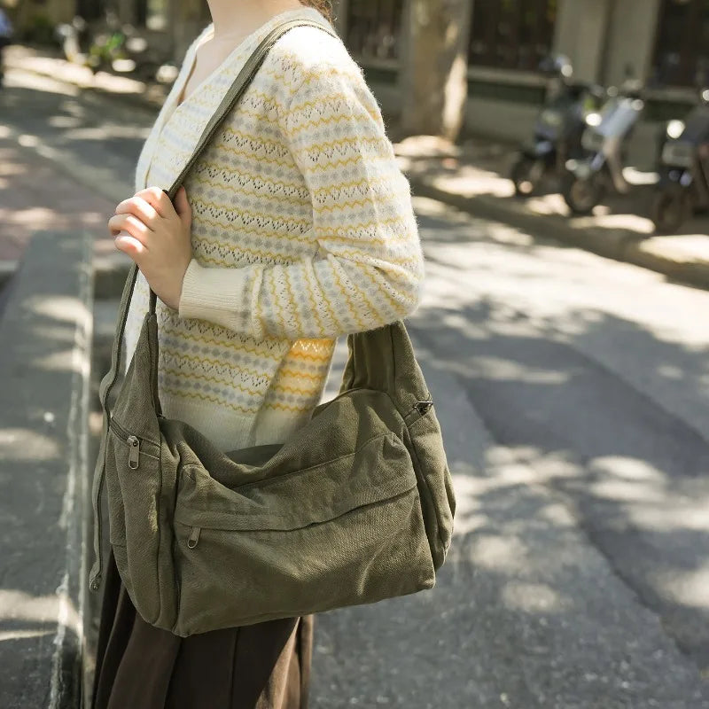 A person wearing a cream and yellow patterned sweater carries a large olive green cotton canvas shoulder crossbody bag on a s