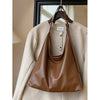 A brown PU leather slouchy hobo shoulder bag is displayed hanging on a wooden hanger over a beige coat.