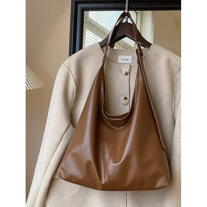 A brown PU leather slouchy hobo shoulder bag is displayed hanging on a wooden hanger over a beige coat.