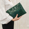 A person holds a dark green alligator print clutch bag against a light background.