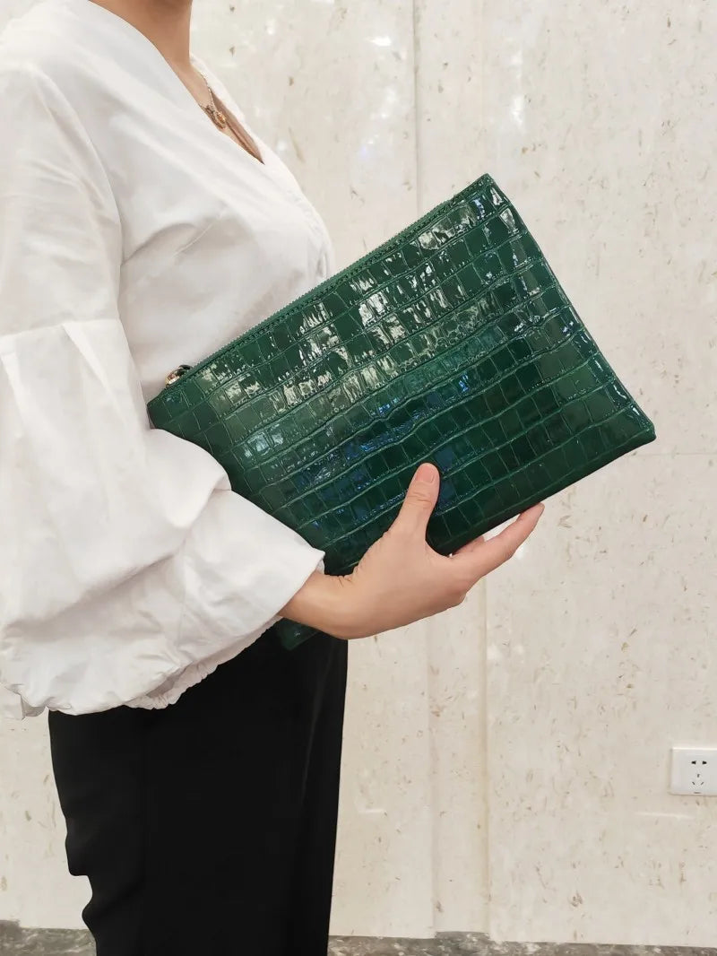 A person holds a dark green alligator print clutch bag against a light background.