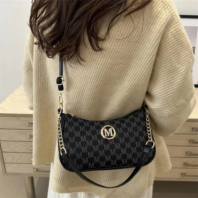 Una manga de suéter beige sostiene un bolso cruz con monograma M en negro y dorado.