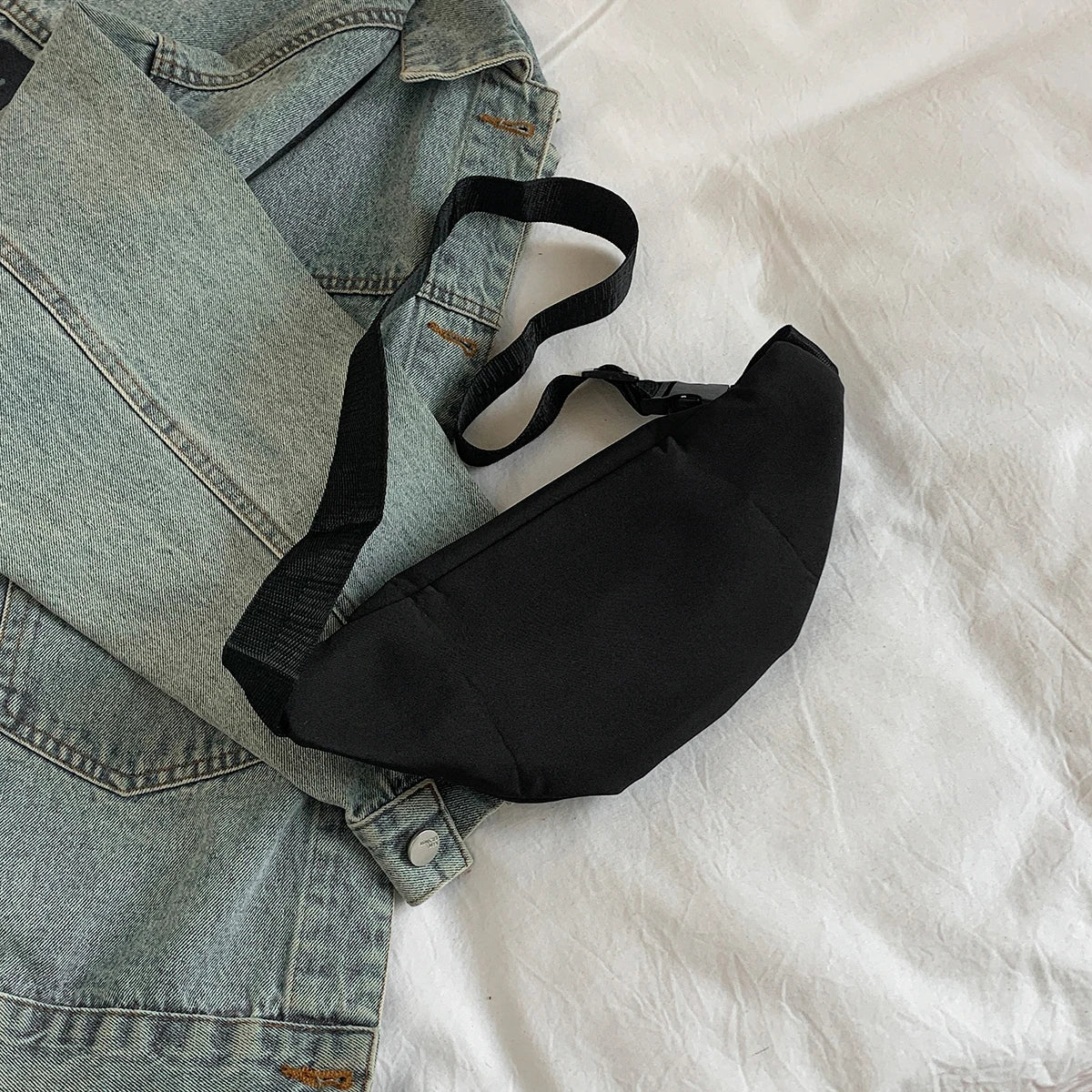 A black nylon crossbody sling bag is shown resting on a light-colored sheet next to a denim shirt.