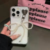 A hand holds a smartphone decorated with a pearl bow keychain and heart pendant against a pink background.