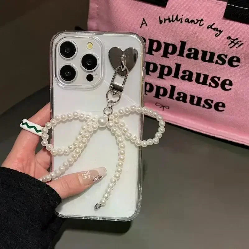 A hand holds a smartphone decorated with a pearl bow keychain and heart pendant against a pink background.
