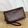Portefeuille pochette en cuir marron foncé avec fermeture éclair et logo gravé, posé sur une surface en bois.