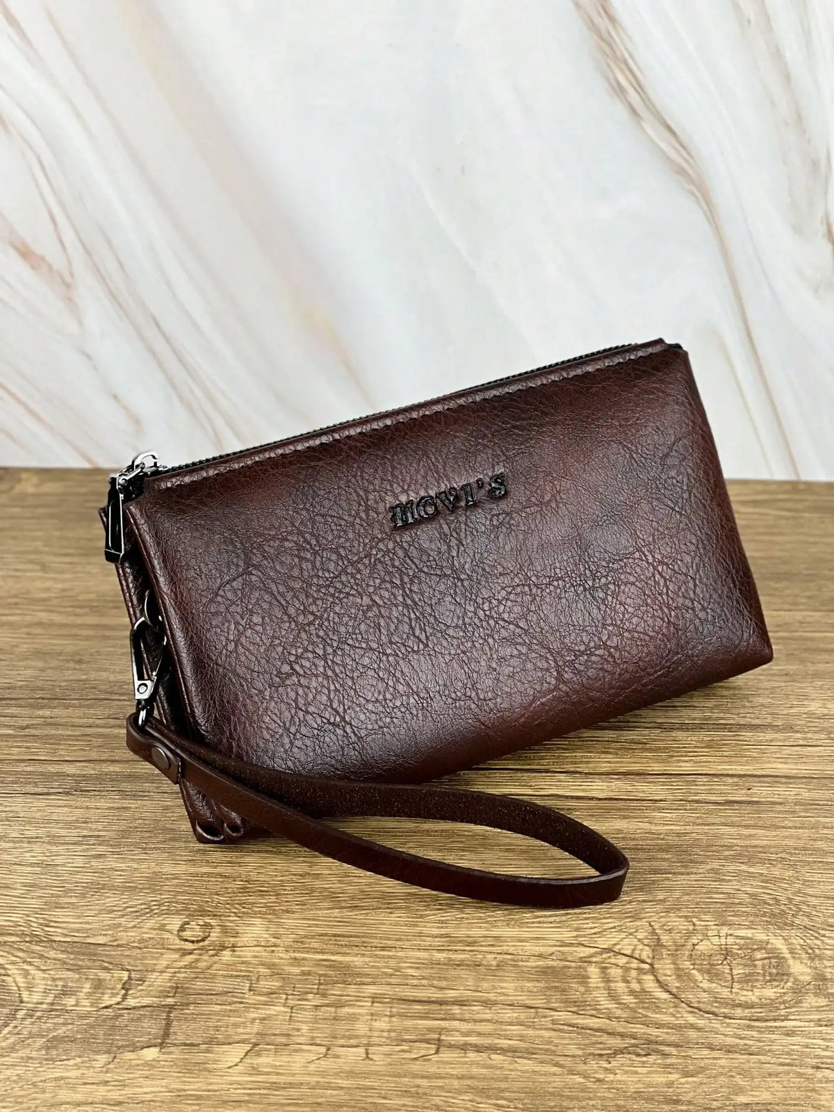 Portefeuille pochette en cuir marron foncé avec fermeture éclair et logo gravé, posé sur une surface en bois.