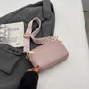 A light pink vegan PU crossbody bag with a patterned strap rests on a gray blazer next to a book.