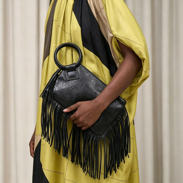A hand holds a black fringed clutch bag with a leather strap against a yellow and black draped garment.